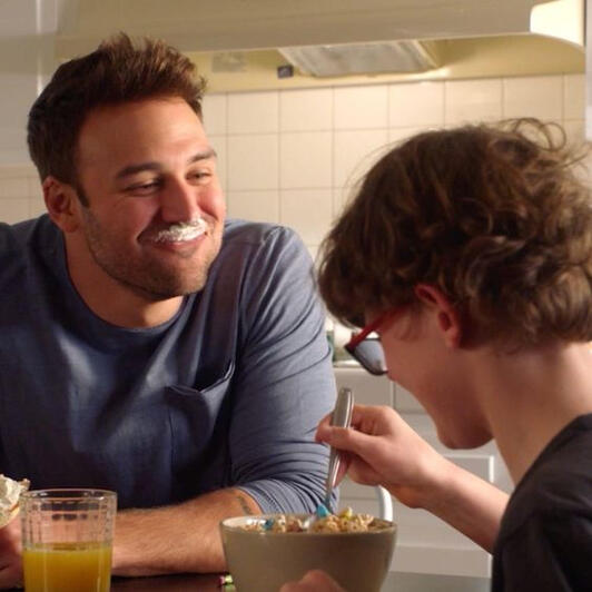 eddie diaz smiling with cream on his upper lip, while his son christopher eats breakfast